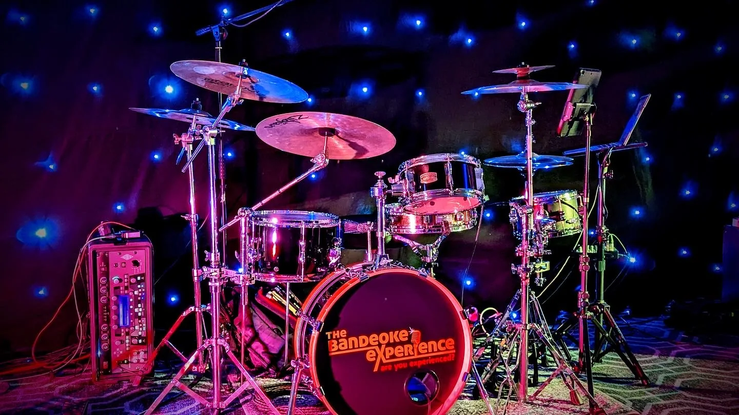 Acoustic drum kit under magenta and blue stage lights against a starcloth backdrop