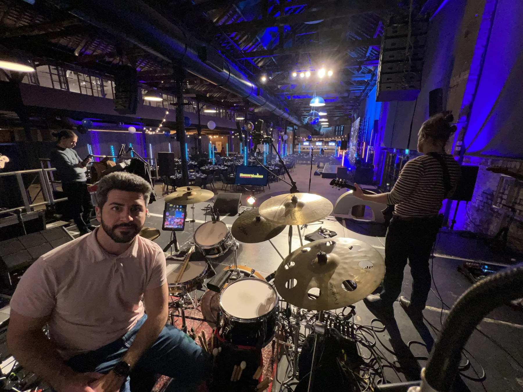 Oscar seated beside his drum kit, with bandmates and an event room behind him